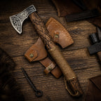 Handmade Viking axe with engraved steel head, carved wood handle, and leather sheath on a workshop table