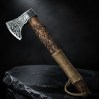 Handmade Viking axe with engraved carbon steel head and carved wood handle standing on a dark stone