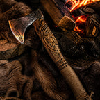 Engraved Viking-style axe resting on fur and leather beside burning firewood, featuring a carved wooden handle and ornate metal axe head.